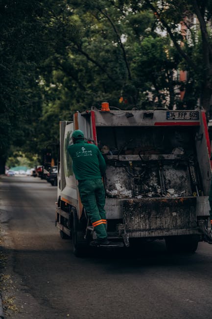 A waste management worker dressed in a green uniform with orange reflective bands is standing next to the open rear compartment of a large, black and white rubbish collection truck on a suburban stree