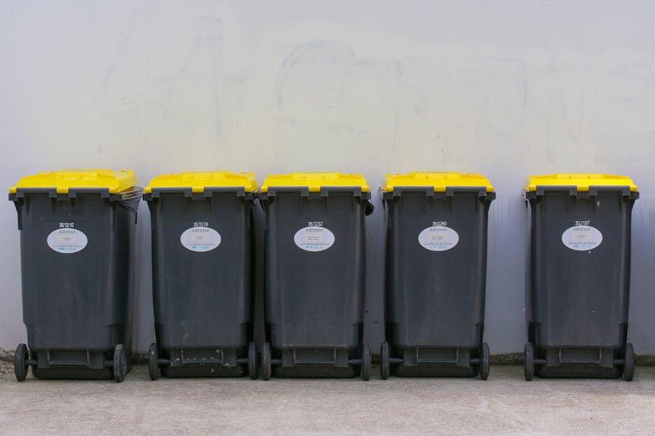 A waste management worker dressed in a green uniform with orange reflective bands is standing next to the open rear compartment of a large, black and white rubbish collection truck on a suburban stree